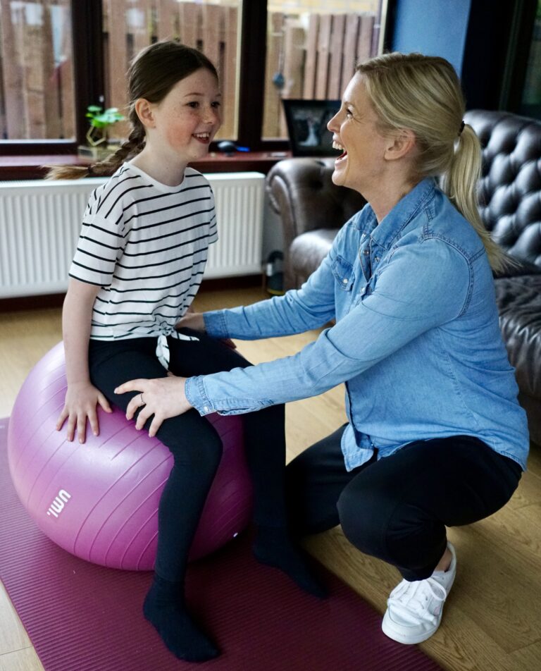 Rachel Moyes, pediatric OT, playfully laughs with a 10-year-old girl sitting on a bright gym ball.